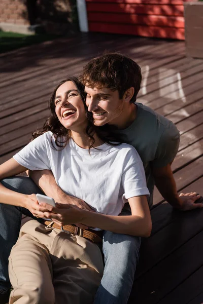Cheerful man hugging brunette girlfriend with smartphone on terrace — Stockfoto
