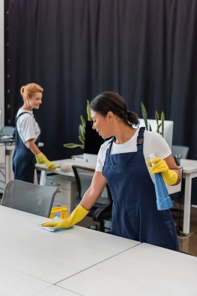 Femme bi-raciale avec détergent et chiffon bureau de nettoyage près de collègue sur fond flou — Photo de stock