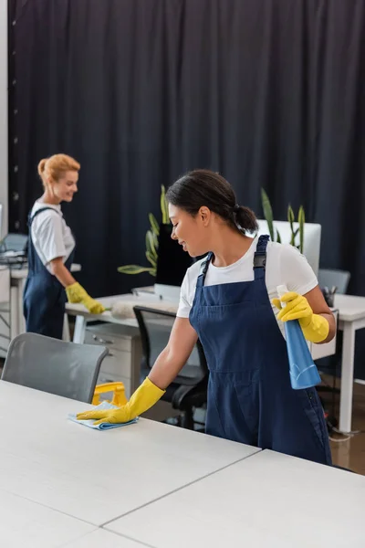 Zwei-Rassen-Frau mit Waschmittel und Putzlappen Büroschreibtisch in der Nähe Kollege auf verschwommenem Hintergrund — Stockfoto