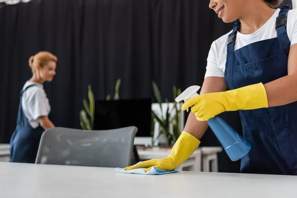 Lächelnde zweirassige Frau putzt Büroschreibtisch mit Waschmittel und Lappen neben verschwommenem Kollegen — Stock Photo