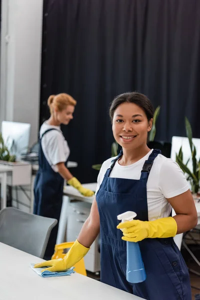 Glückliche zweirassige Frau in Overalls und Gummihandschuhen, die in die Kamera neben verschwommenem Kollegen lächelt — Stockfoto