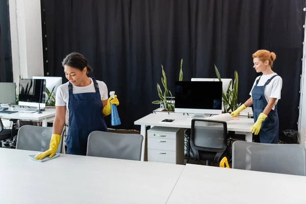 Donne interrazziali con stracci e spazzole antipolvere scrivanie di pulizia in ufficio — Foto stock