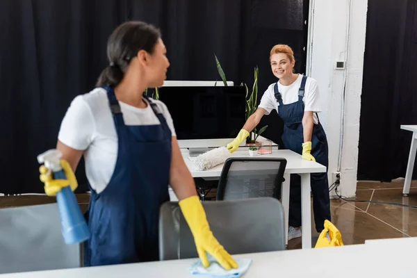 Femme souriante avec brosse à poussière regardant un collègue bi-racial au premier plan flou — Photo de stock