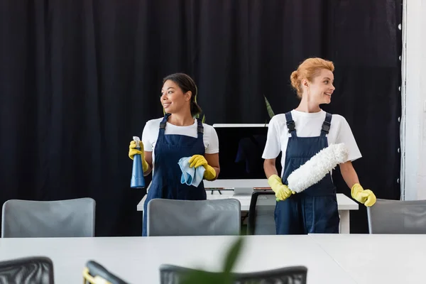 Interracial women in overalls holding cleaning supplies and smiling in office — Stock Photo