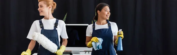 Cheerful multiethnic cleaners holding dust brush and rag in office, banner — Stock Photo
