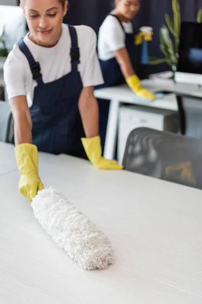 Lächelnde Frau putzt Schreibtisch mit Staubbürste neben verschwommenem birassischen Kollegen — Stock Photo