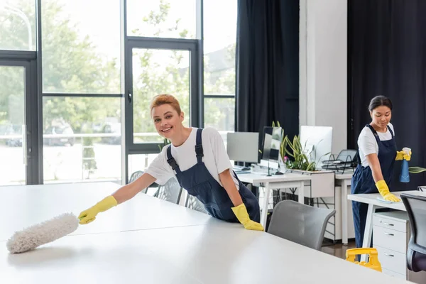 Happy woman in overalls cleaning office desk with dust brush near bi-racial colleague on background — Stock Photo