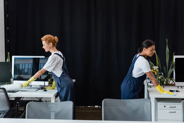 Side view of multiethnic women cleaning office near computer monitors with blank screen — Stock Photo