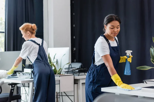 Interracial femmes en salopette nettoyage bureau avec brosse à poussière et chiffon — Photo de stock