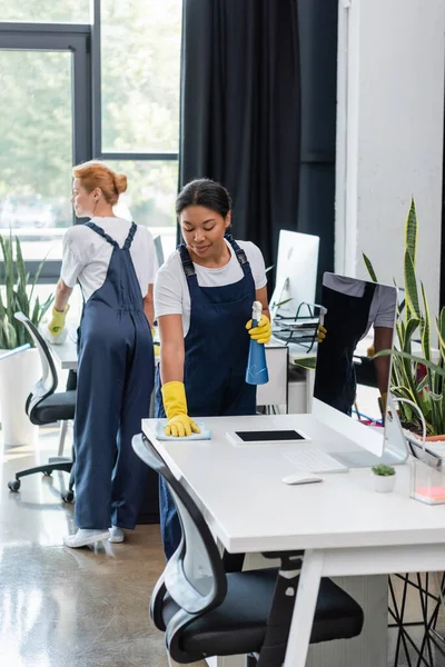 Mulheres multiculturais em mesas de limpeza uniformes perto de monitores de computador no escritório — Fotografia de Stock