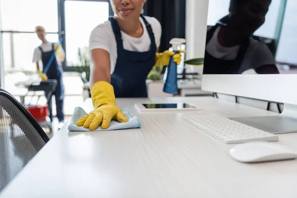 Vista ritagliata della donna bi-razziale in guanti di gomma ufficio di pulizia scrivania vicino gadget e collega su sfondo sfocato — Foto stock