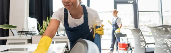 Mujer bi-racial sonriente con oficina de limpieza de detergente cerca de colega borrosa, pancarta - foto de stock