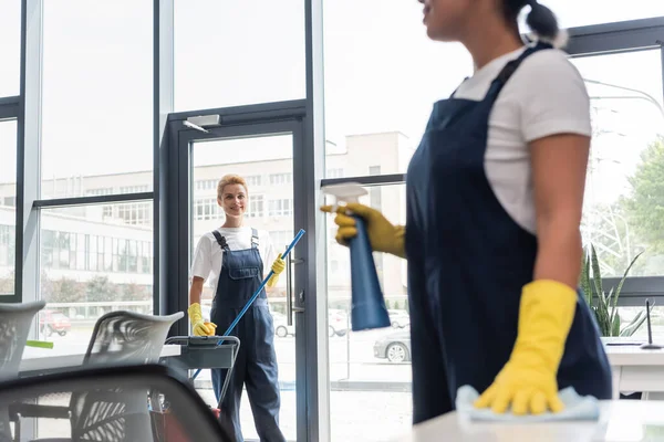 Femme souriante en salopette regardant un collègue bi-racial avec du détergent sur le premier plan flou — Photo de stock