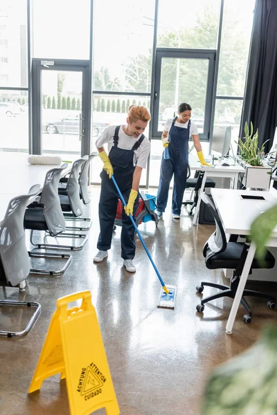 Femme heureuse plancher de lavage avec balai près du conseil de prudence et collègue bi-raciale — Photo de stock