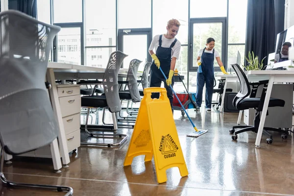 Femme en salopette plancher de lavage avec balai près du conseil d'attention et collègue bi-raciale — Photo de stock