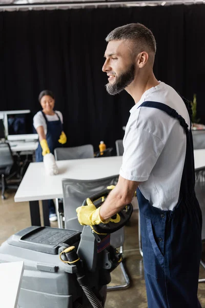 Bärtiger Mann mit Bodenwaschmaschine in der Nähe einer Frau, die auf verschwommenem Hintergrund Schreibtisch wischt — Stockfoto