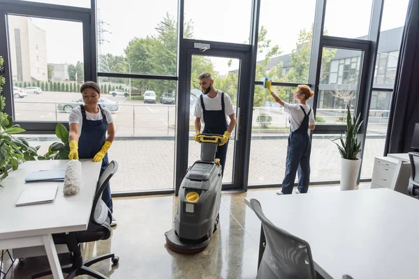 Man with floor scrubber machine near interracial women washing windows and furniture — Stock Photo