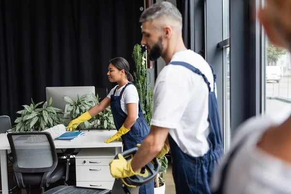 Vue latérale de l'homme avec machine de nettoyage de sol et bureau de nettoyage de femme bi-raciale au premier plan flou — Photo de stock