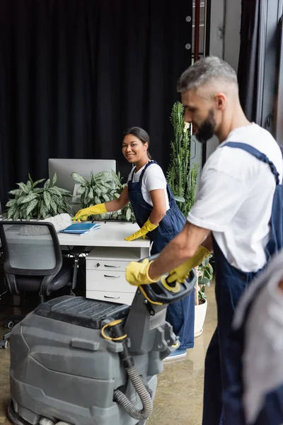 Bi-racial mujer sonriendo a la cámara cerca de hombre con piso máquina de lavado en primer plano borrosa - foto de stock