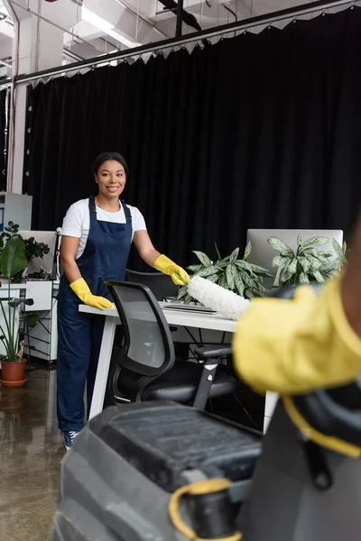 Junge und lächelnde zweirassige Frau mit Staubbürste, die Büroschreibtisch neben Mann auf verschwommenem Vordergrund putzt — Stockfoto