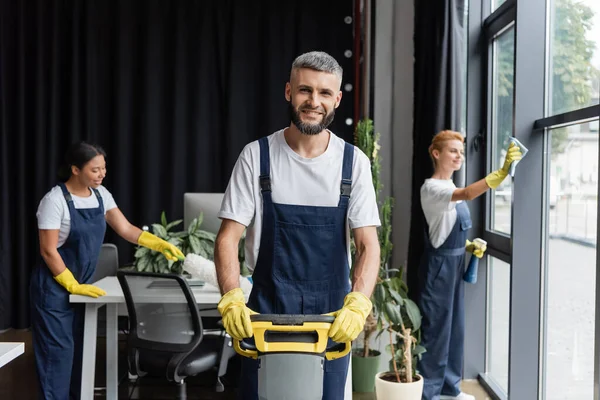 Homem feliz com máquina de limpeza de piso olhando para a câmera perto de mulheres multiétnicas escritório de limpeza — Fotografia de Stock