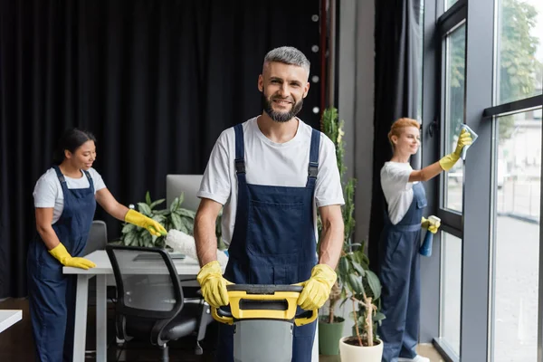 Homme heureux avec laveur de sol machine regardant la caméra près de femmes multiethniques bureau de nettoyage — Photo de stock