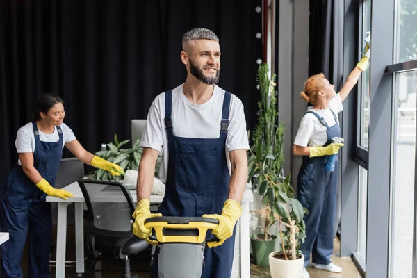 Bärtiger Mann mit Bodenwaschmaschine, der in der Nähe eines interrassischen Teams, das am Hintergrund arbeitet, wegschaut — Stockfoto