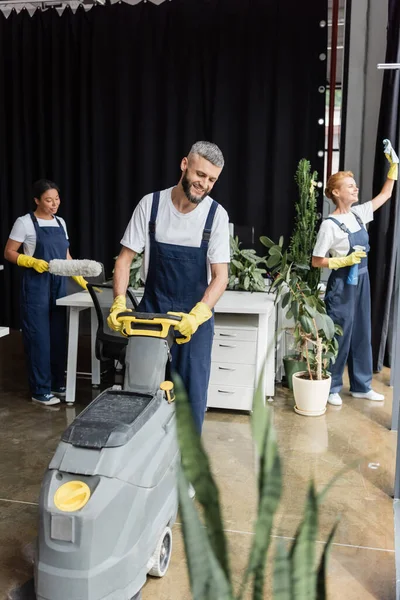Felice uomo funzionamento lavasciuga pavimenti macchina mentre le donne multietniche pulizia ufficio — Foto stock