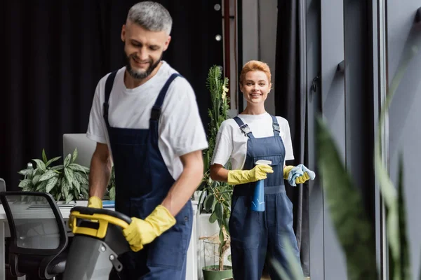 Happy woman with detergent and rag looking at camera near man working on blurred foreground — Stock Photo