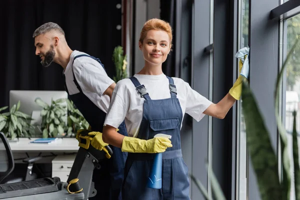 Mujer feliz lavando las ventanas de la oficina cerca del hombre con la máquina eléctrica del depurador del piso - foto de stock