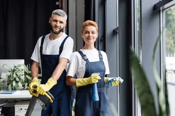 Limpiadores profesionales sonrientes en uniforme mirando la cámara en la oficina - foto de stock