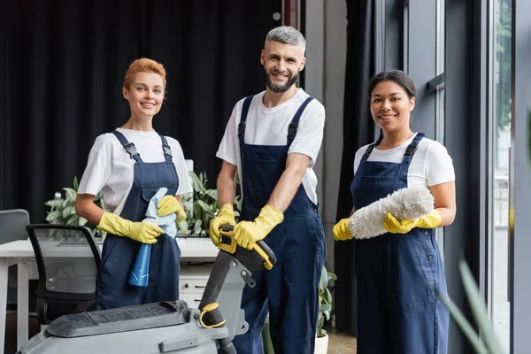 Felice squadra multietnica in abbigliamento da lavoro guardando la fotocamera vicino alle forniture di pulizia — Foto stock