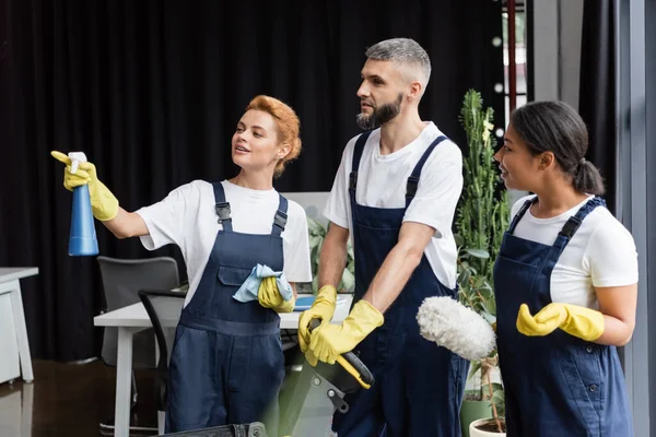 Woman with detergent pointing with finger near interracial colleagues with cleaning supplies — Stock Photo