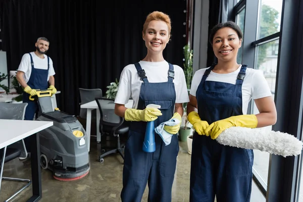 Glückliche interrassische Frauen in Overalls, die in die Kamera in der Nähe des Mitarbeiters schauen, der im Hintergrund den Fußboden reinigt — Stockfoto