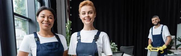 Cheerful interracial women in overalls looking at camera near colleague on blurred background, banner — Stock Photo