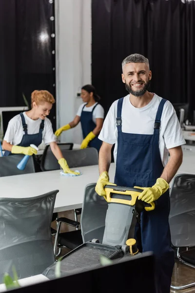 Hombre barbudo alegre con máquina de fregar piso cerca de la oficina de limpieza de las mujeres multiculturales sobre fondo borroso - foto de stock