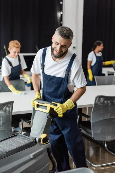 Homme barbu souriant avec machine de nettoyage de sol travaillant près des femmes interracial sur fond flou — Photo de stock