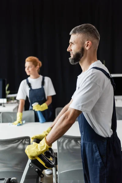 Bärtiger Mann bedient Fußbodenwaschmaschine in der Nähe von Frau, die auf verschwommenem Hintergrund arbeitet — Stockfoto