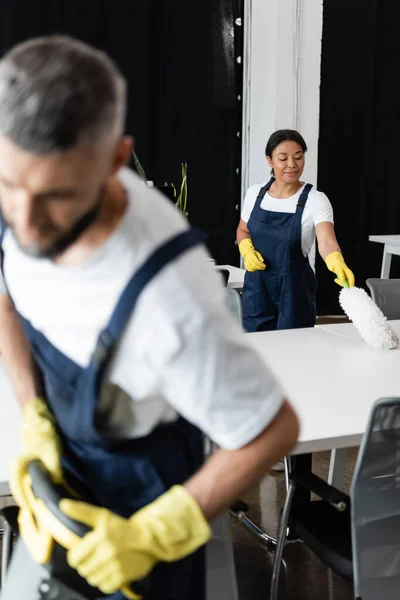 Bi-racial woman in overalls and rubber gloves cleaning desk with dust brush on blurred foreground — Stock Photo