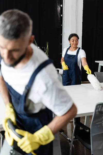 Mujer bi-racial en overoles y guantes de goma escritorio de limpieza con cepillo de polvo en primer plano borroso - foto de stock