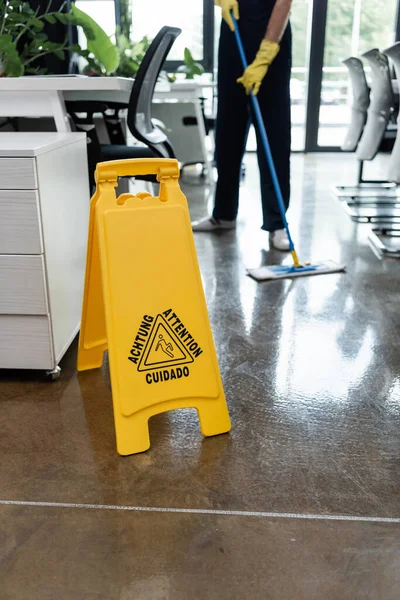 Selective focus of caution board near cropped man washing floor with mop on blurred background — Stock Photo