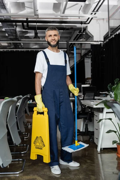 Full length of man in overalls holding mop and attention sign board while smiling at camera — Stock Photo