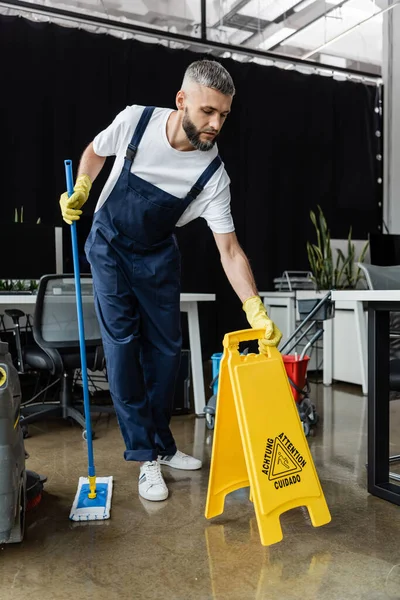 Professional cleaner in overalls holding mop and caution sign board in office — Stock Photo