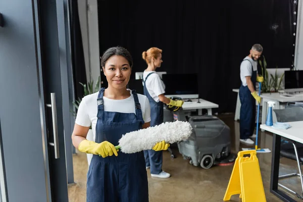 Giovane donna bi-razziale in tuta da lavoro con spazzola antipolvere e guardando la fotocamera vicino ai colleghi di lavoro — Foto stock