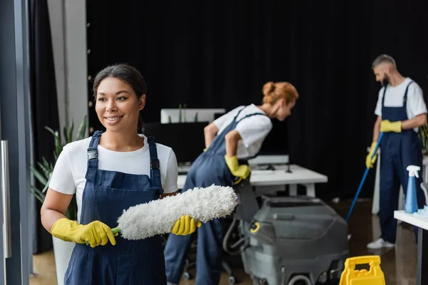 Lächelnde Frau mit Staubbürste in der Nähe von Kollegen und Bodenwaschmaschine auf verschwommenem Hintergrund — Stockfoto