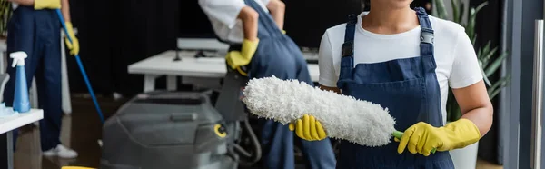 Abgeschnittene Ansicht einer zweirassigen Frau in Gummihandschuhen mit Staubbürste in der Nähe von Kollegen auf verschwommenem Hintergrund, Banner — Stockfoto