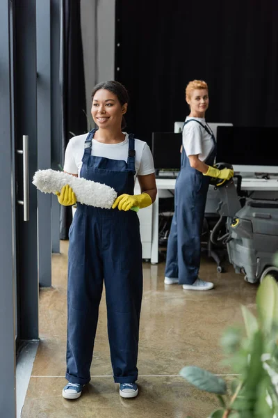 Alegre bi-racial mujer en ropa de trabajo de pie con cepillo de polvo cerca de colega con máquina de fregar piso - foto de stock