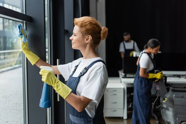 Frau in Gummihandschuhen putzt Fenster mit Lappen und Spülmittel neben verschwommenen interrassischen Kollegen — Stockfoto