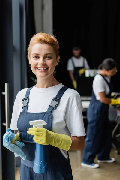 Cheerful woman with rag and spray bottle smiling at camera while colleagues working on blurred background — Stock Photo