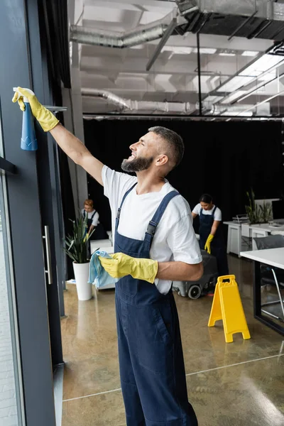 Bearded man in workwear cleaning window with detergent near multiethnic women working in office — Stock Photo